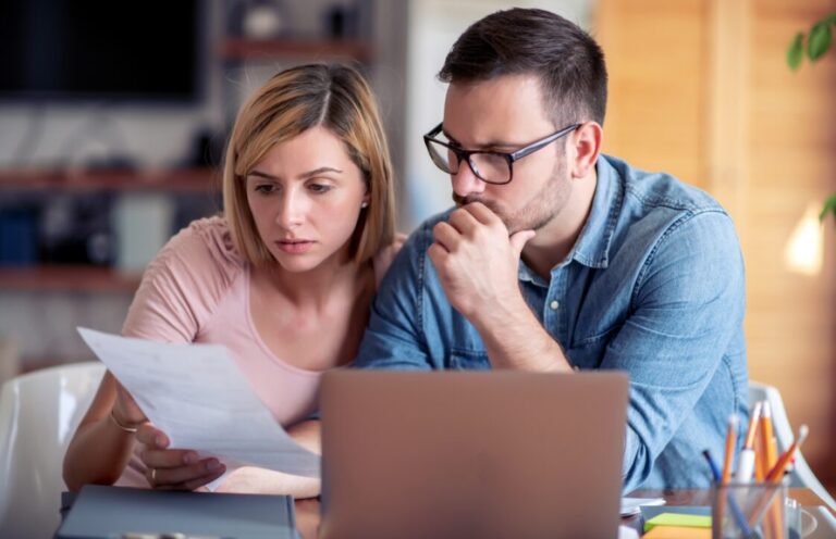 A couple sits at a table, looking intently at a piece of paper, with a laptop and pens nearby.